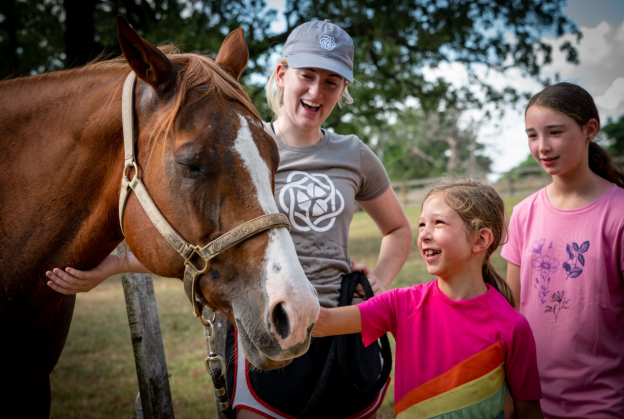 Three girls smile while interacting with a horse